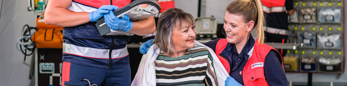 Eine ehrenamtliche Sanitäterin legt einer Dame eine Decke um die Schulter. Im Hintergrund schreibt ein Sanitäter Notizen auf ein Klemmbrett.